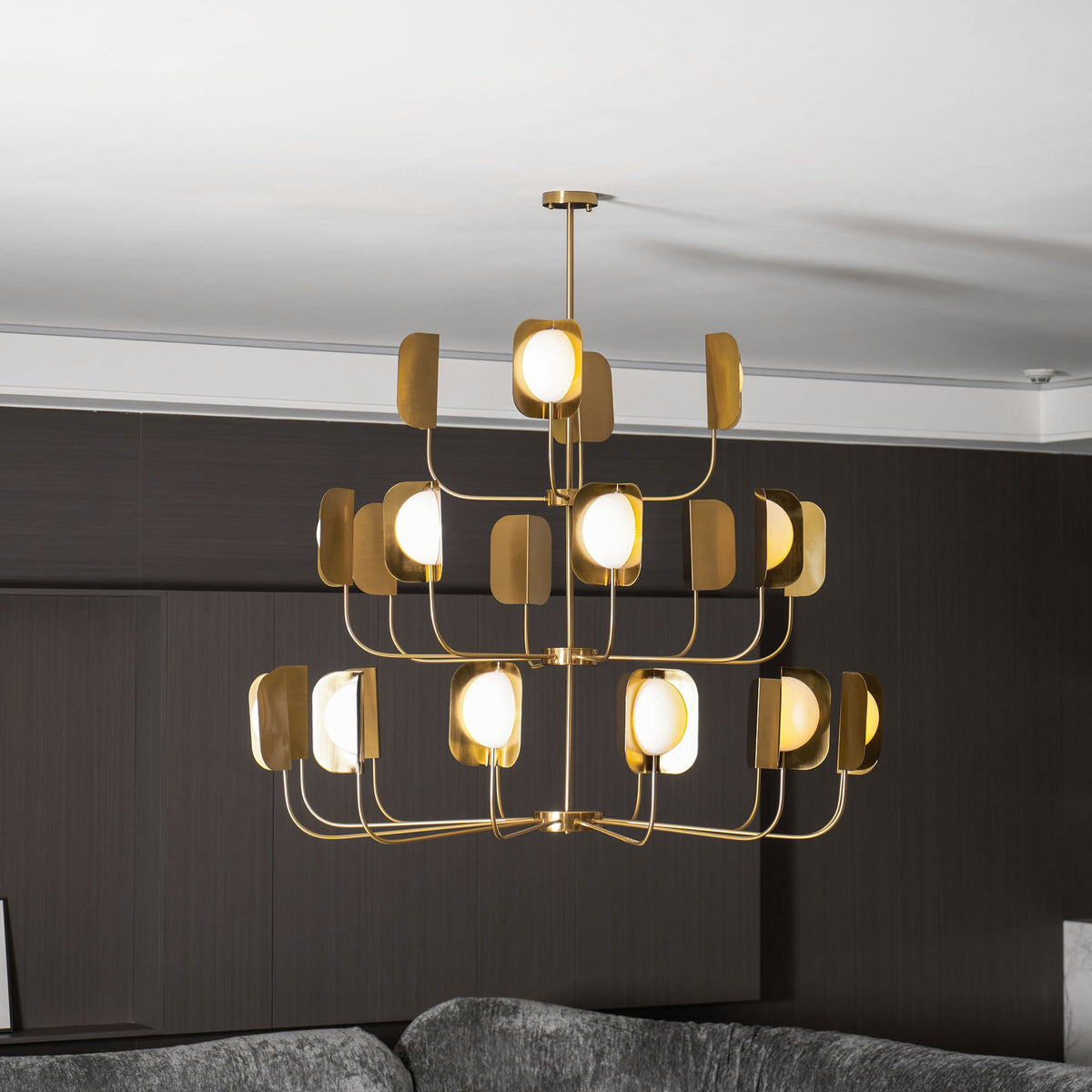 Three tiers of lit leaf shades on a brass-toned Clérachic chandelier, seen straight on above a sofa in a dark panelled room.