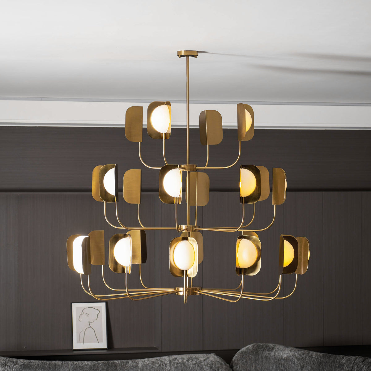 The lit brass-toned Clérachic chandelier seen from the side, its lower tier of arms spread wide above a low table and a small framed print.