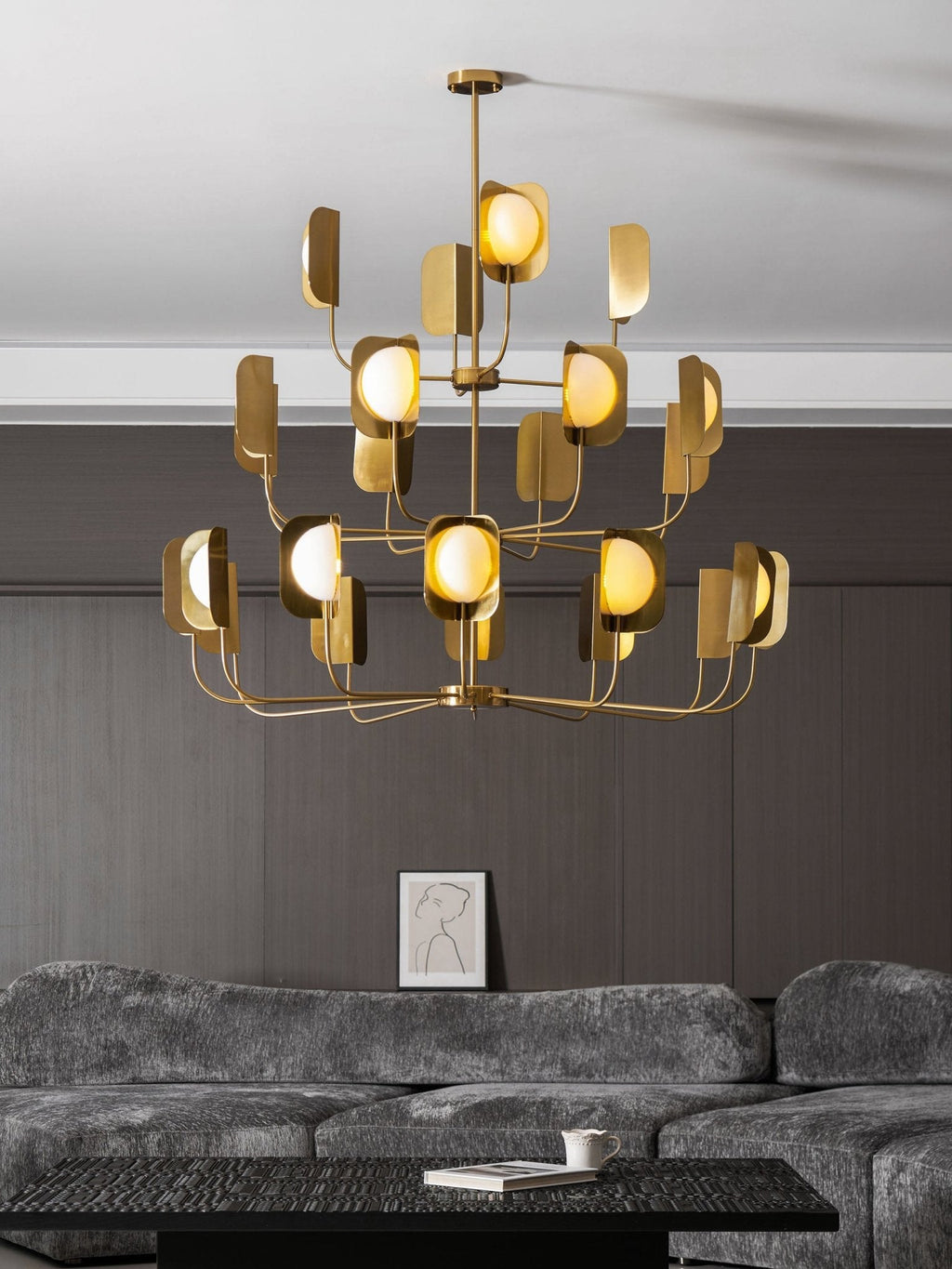 Low view of a brass-toned Clérachic chandelier in a lounge, its three tiers of leaf shades and globes against a dark wall.