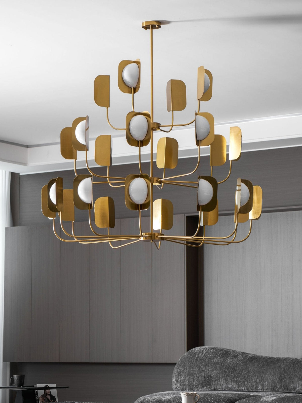 A brass-toned Clérachic chandelier hung in front of a grey wall with a sofa below, its three tiers of leaf shades and globes lit.