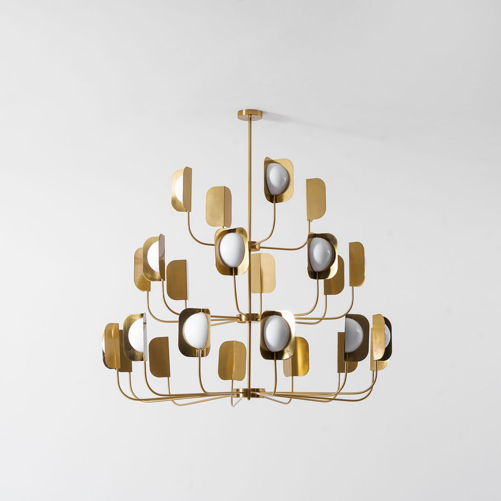 Unlit, this brass-toned Clérachic chandelier shows three tiers of leaf shades and white globes against a pale grey wall.