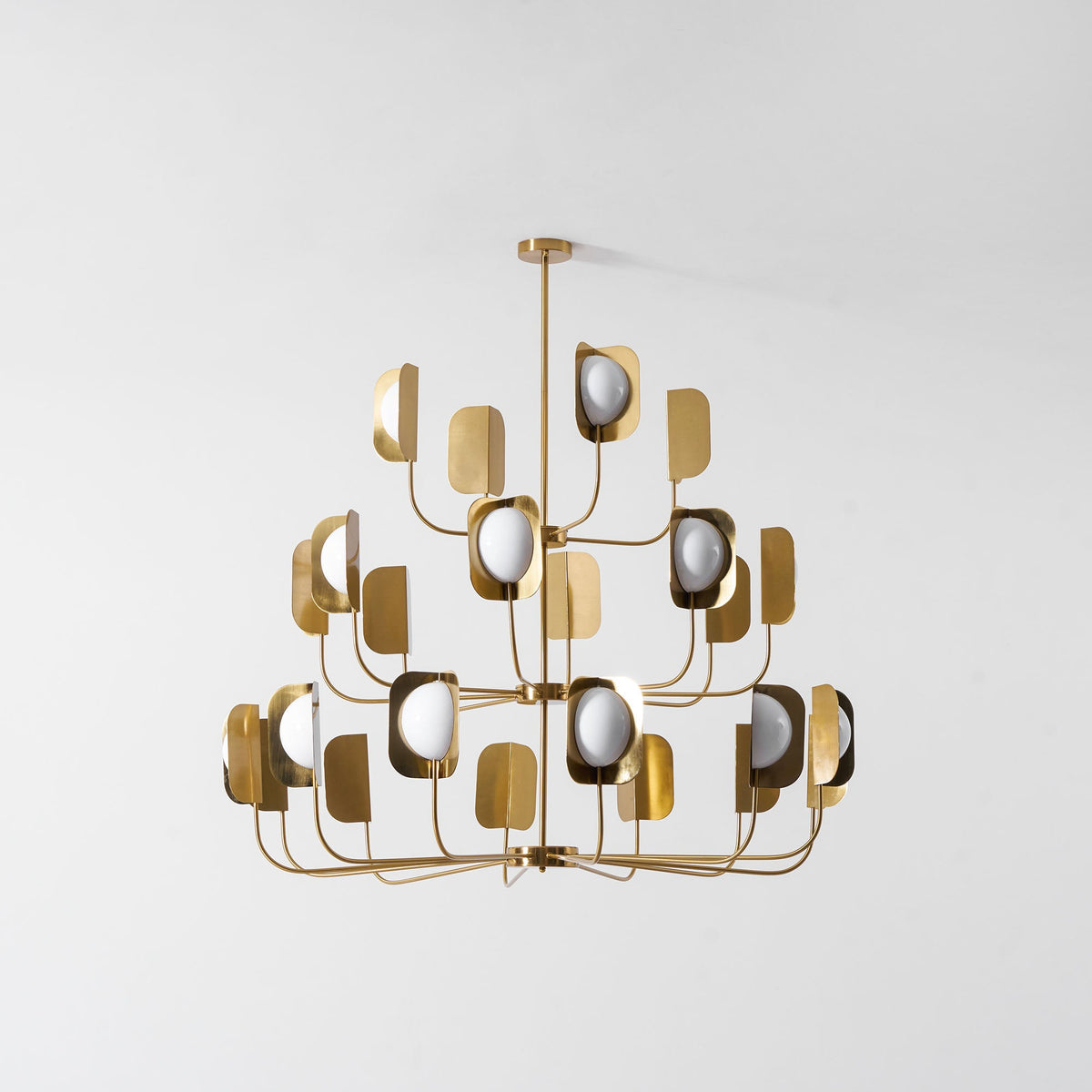 Unlit, this brass-toned Clérachic chandelier shows three tiers of leaf shades and white globes against a pale grey wall.