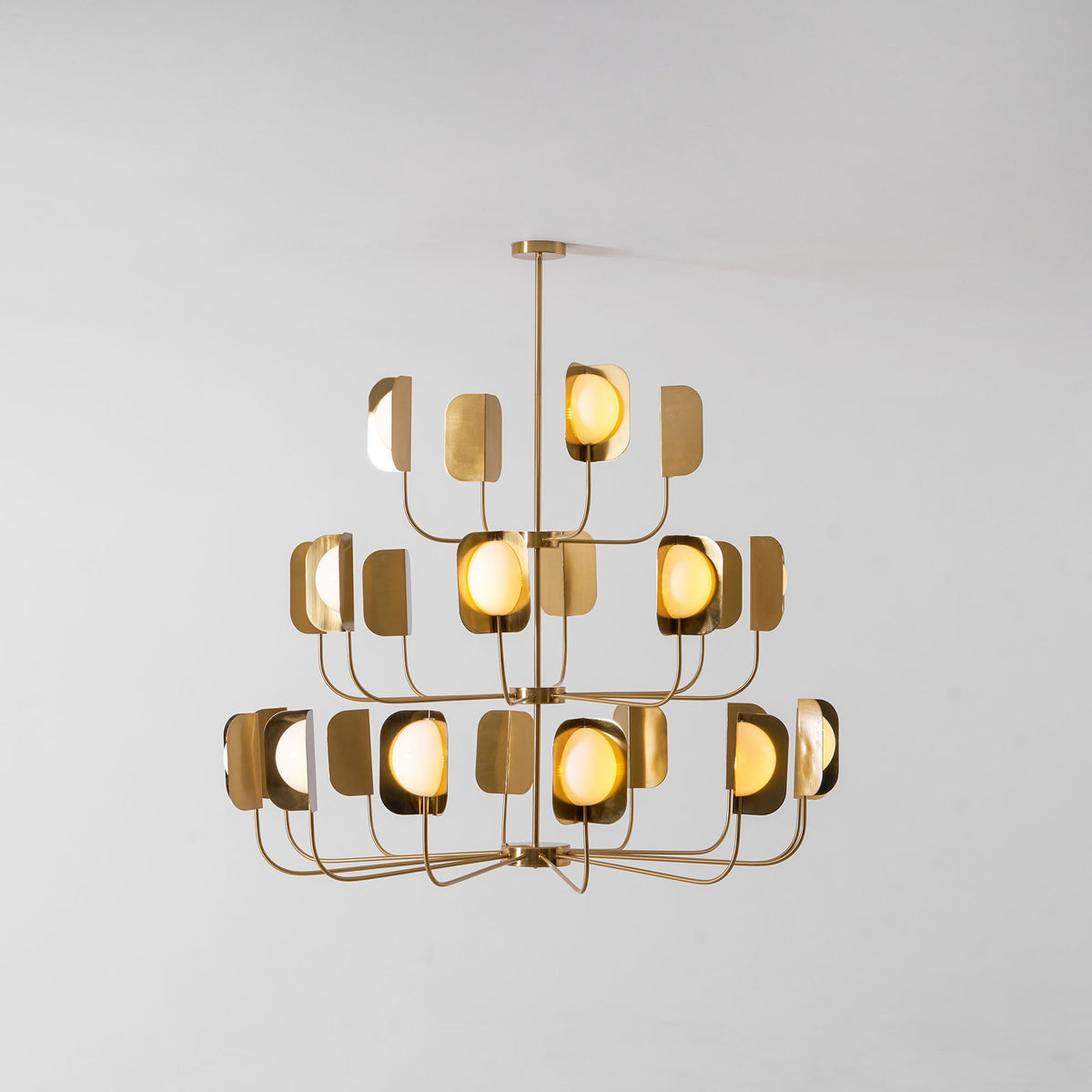 A brass-toned Clérachic chandelier lit warm on a pale backdrop, each globe glowing inside its leaf shade on three tiers.