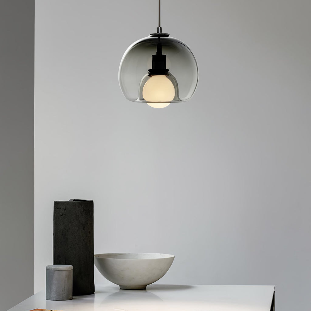 A smoke glass Clérachic pendant hangs in a pale room above a stone bowl and a dark vase on a table edge.