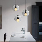 Three Clérachic pendants in clear glass hang at staggered heights above a white table, with a dark door behind.