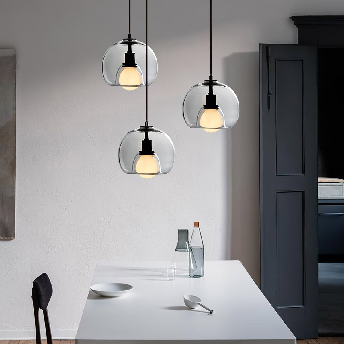 Three Clérachic pendants in clear glass hang at staggered heights above a white table, with a dark door behind.