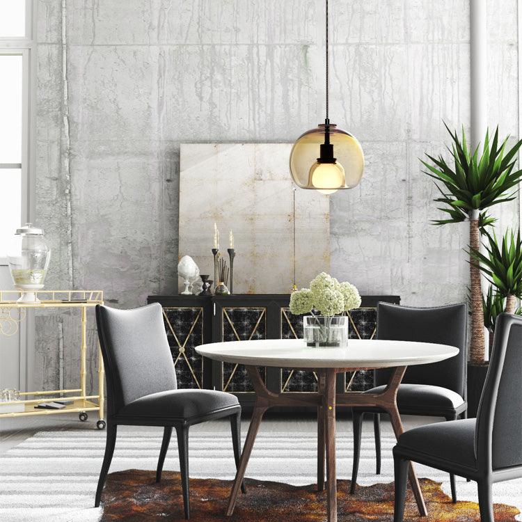 A cognac glass Clérachic pendant hangs over a round white table and black chairs in front of a raw concrete wall.