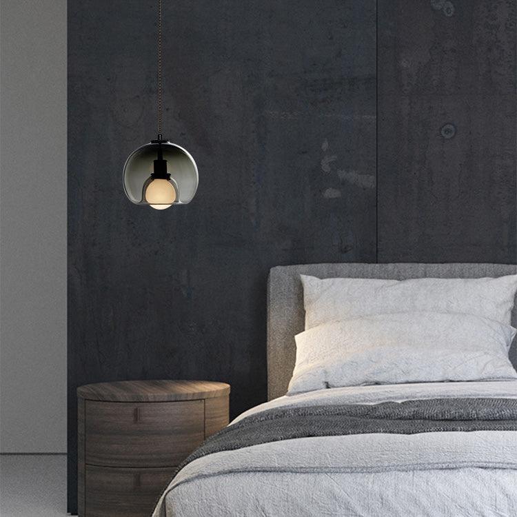 A smoke glass Clérachic pendant hangs above a bedside table and grey bed, in front of a dark textured wall.