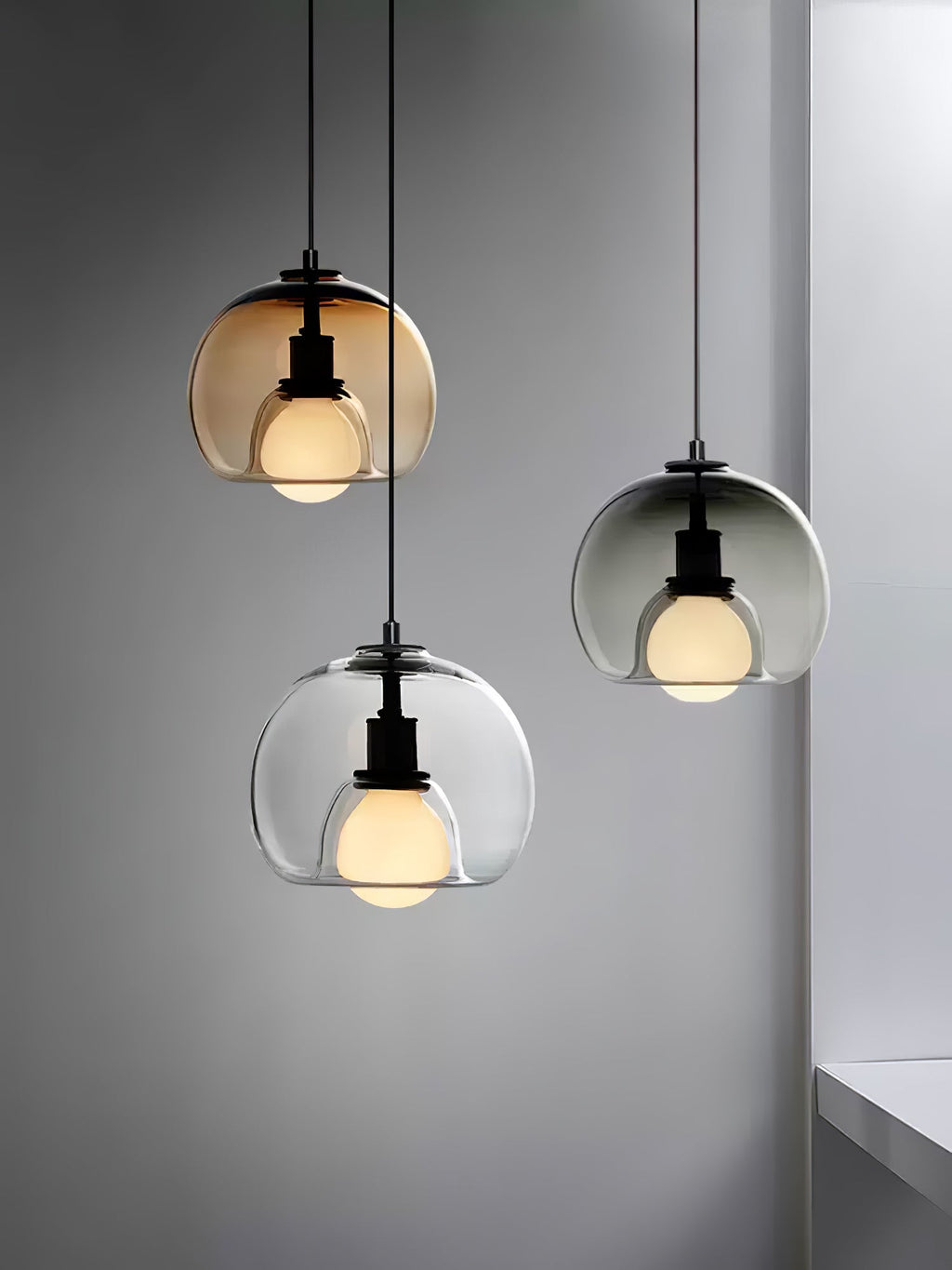Three Clérachic pendants in cognac, clear and smoke glass hang at staggered heights against a grey wall in a tall frame.