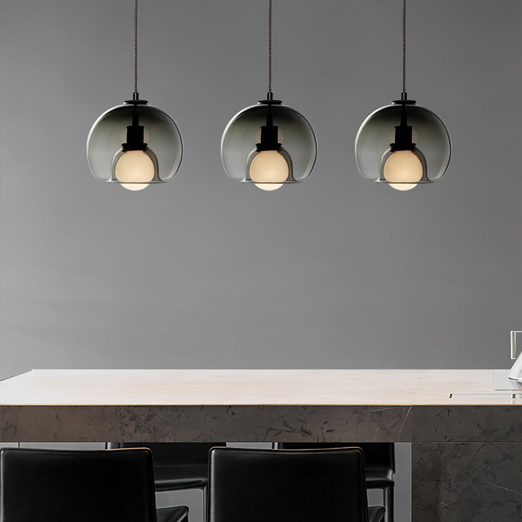 Three smoke glass Clérachic pendants hang in a row over a pale stone counter with black stools in a grey room.