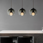 Three smoke glass Clérachic pendants hang in a row over a pale stone counter with black stools in a grey room.