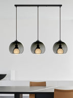 A Clérachic trio in smoke glass hangs from one black bar canopy over a dark table with tan chairs in a white room.