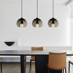 Three smoke glass Clérachic pendants on a black bar canopy hang above a dark table with tan leather chairs.