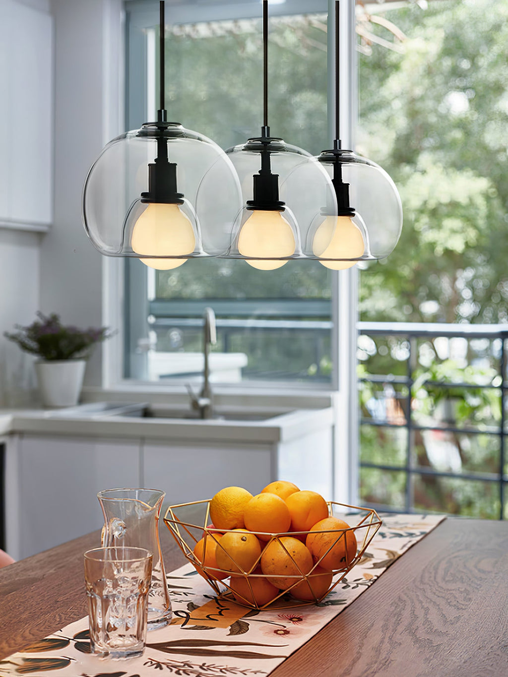 Three clear glass Clérachic pendants hang close together over a wood table with an orange bowl and a window behind.