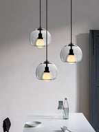 Three clear glass Clérachic pendants hang at different heights above a white table, beside a tall dark door.