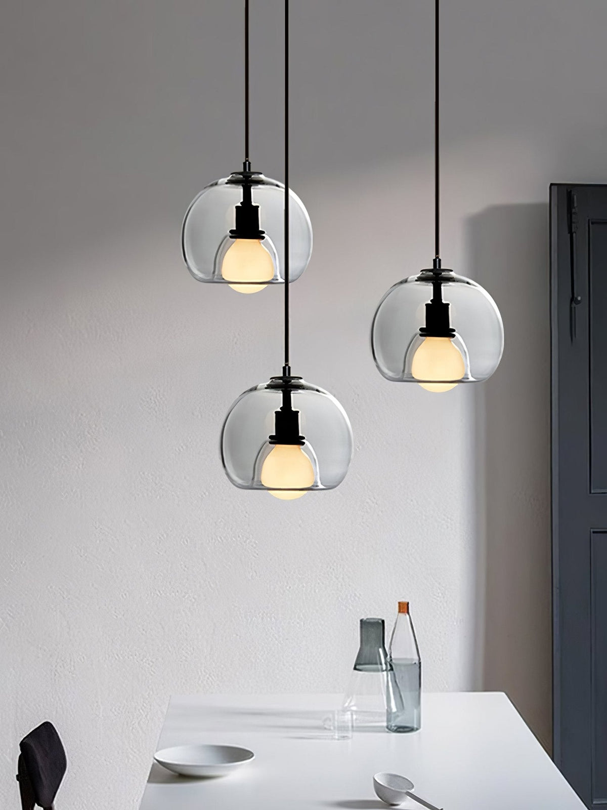 Three clear glass Clérachic pendants hang at different heights above a white table, beside a tall dark door.