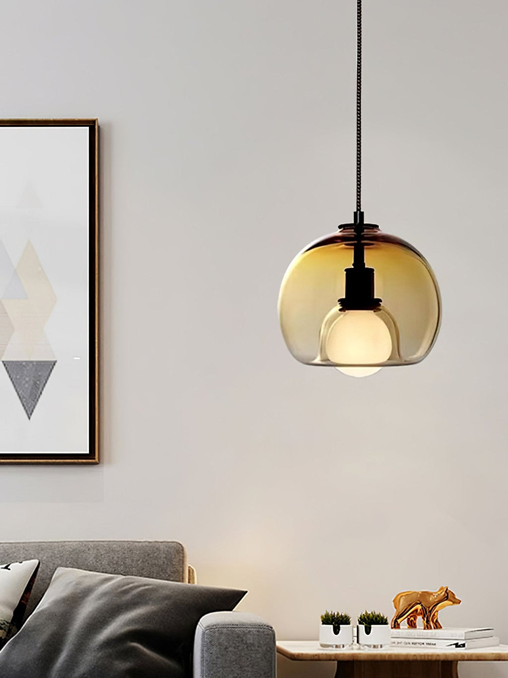 A cognac glass Clérachic pendant hangs by a framed geometric print, with a grey sofa and cushions below.