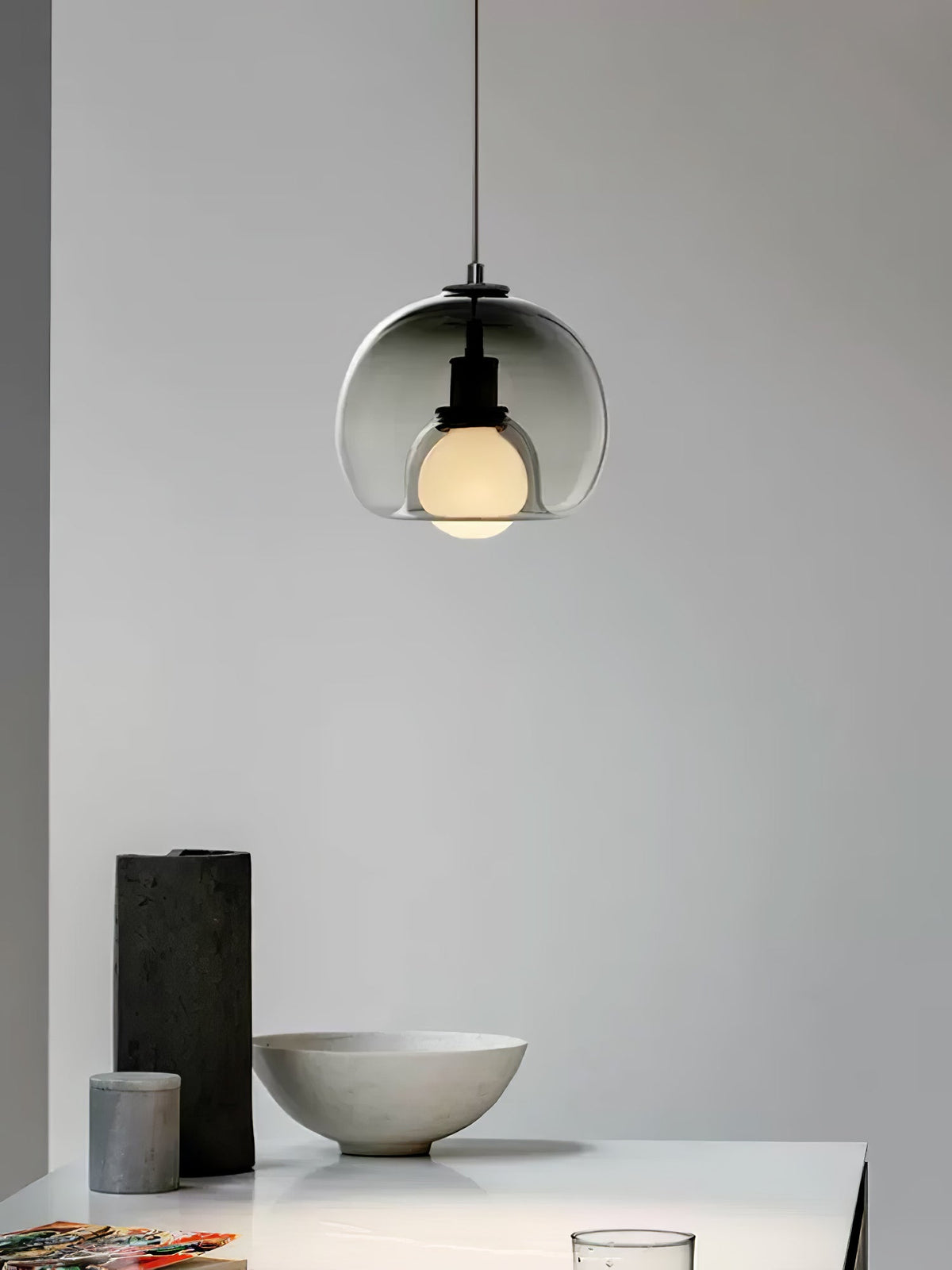A smoke glass Clérachic pendant hangs in a pale room, with a black vase and stone bowl on the table below.