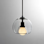 A clear glass Clérachic pendant on a black cord, shown on a grey gradient backdrop with its frosted bulb lit.