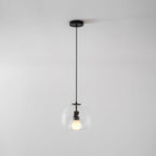 A clear glass Clérachic pendant with a bulb lit hangs from a round black canopy against a plain grey wall.