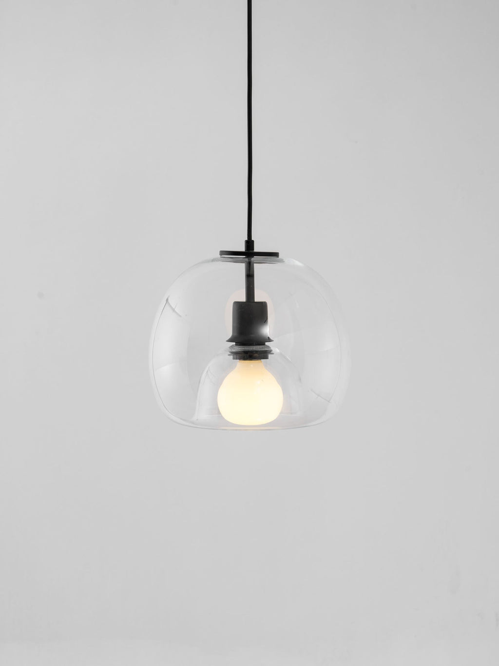 A clear glass Clérachic pendant hung from a black cord against a pale grey backdrop, with a lit frosted bulb.