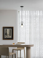 A clear glass Clérachic pendant hangs in a bright room in front of white patterned curtains, above a pale table.