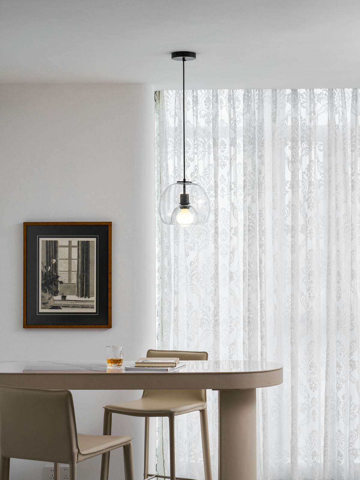 A clear glass Clérachic pendant hangs in a bright room in front of white patterned curtains, above a pale table.
