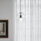 A Clérachic pendant in clear glass hangs by a framed print and sheer patterned curtains, with a table edge below.
