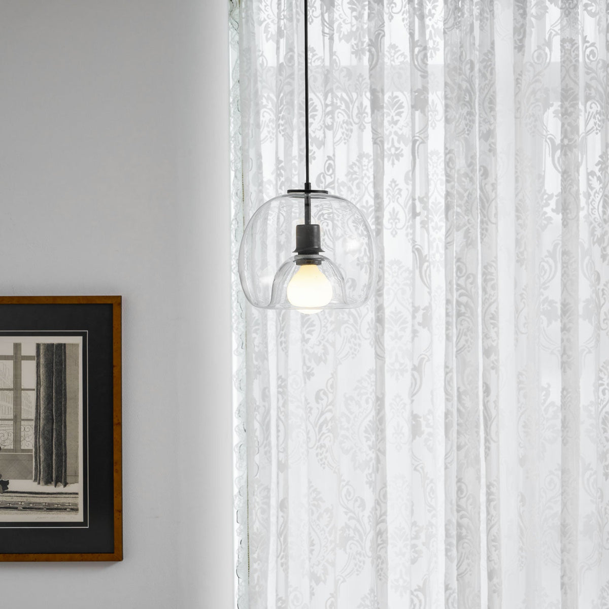 A Clérachic pendant in clear glass hangs by a framed print and sheer patterned curtains, with a table edge below.