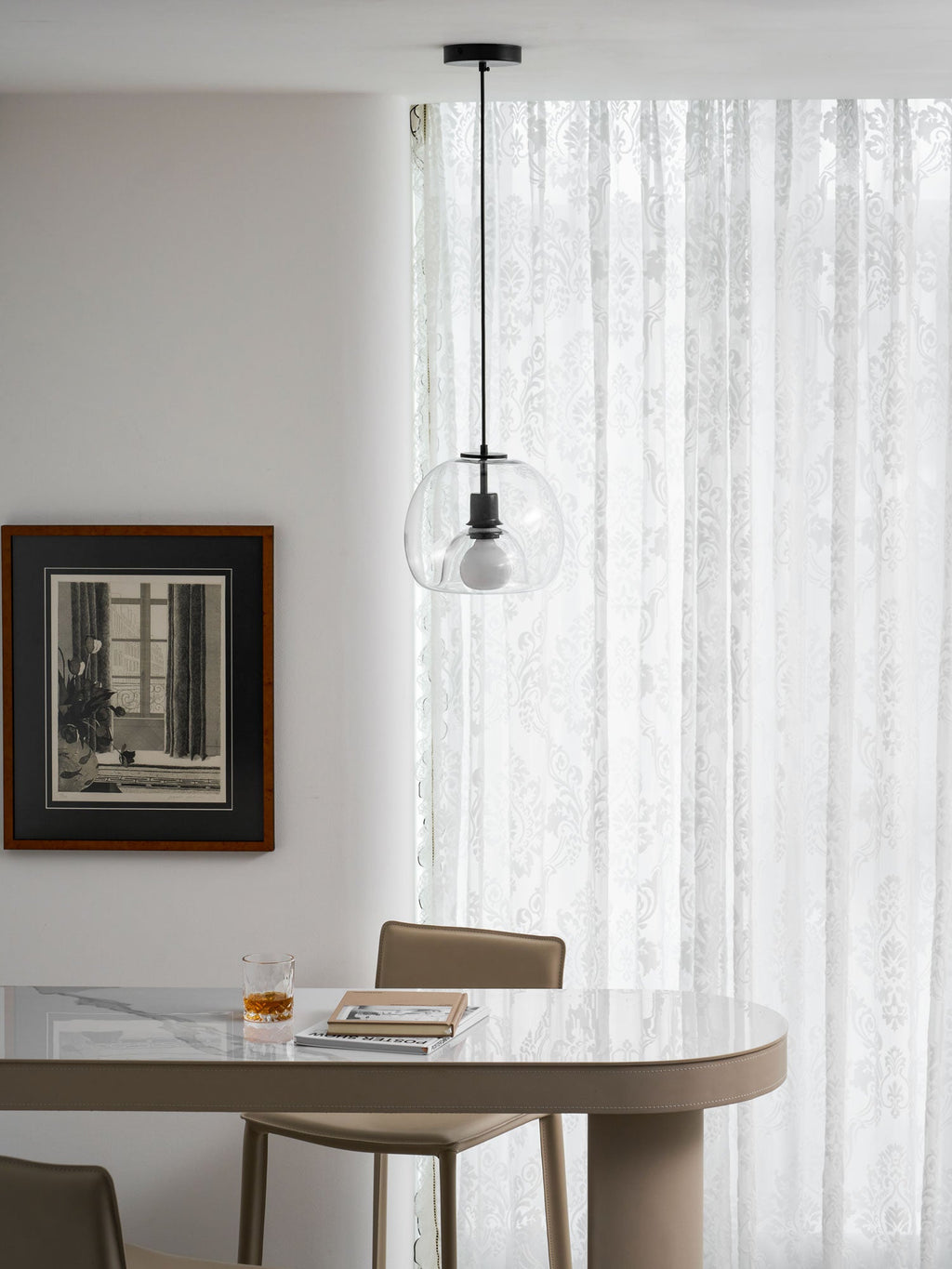 A small clear Clérachic pendant hangs in front of sheer patterned curtains, above a table with a glass and books.