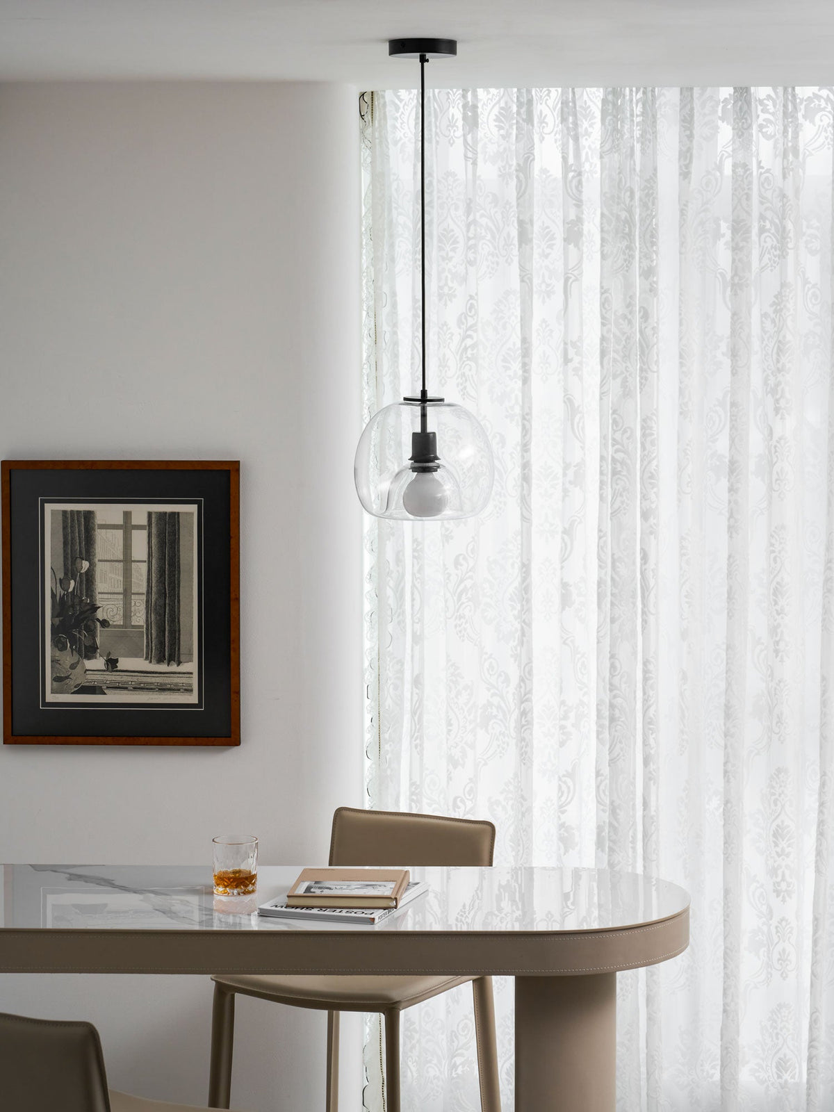 A small clear Clérachic pendant hangs in front of sheer patterned curtains, above a table with a glass and books.