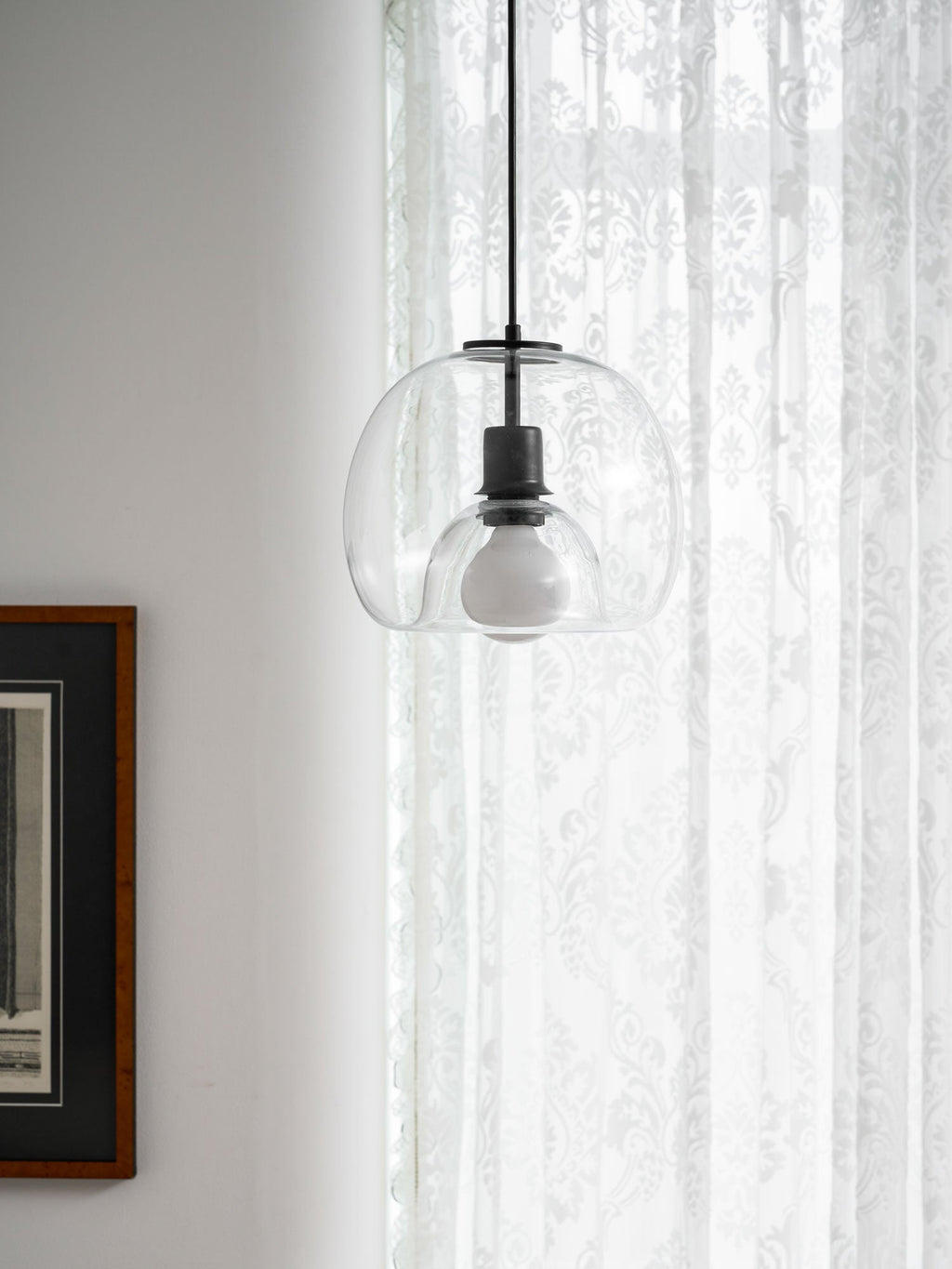 A clear glass Clérachic pendant glows softly against lace curtains, beside a dark framed print.