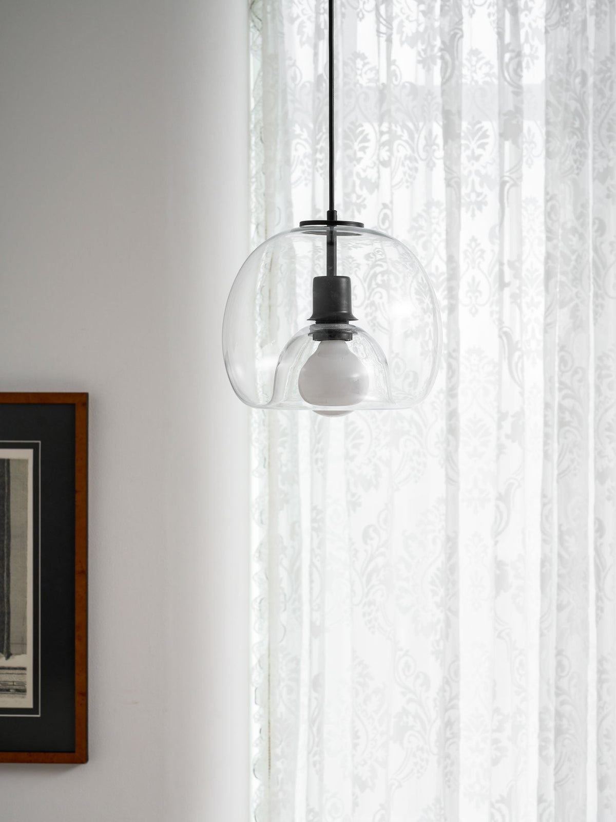 A clear glass Clérachic pendant glows softly against lace curtains, beside a dark framed print.