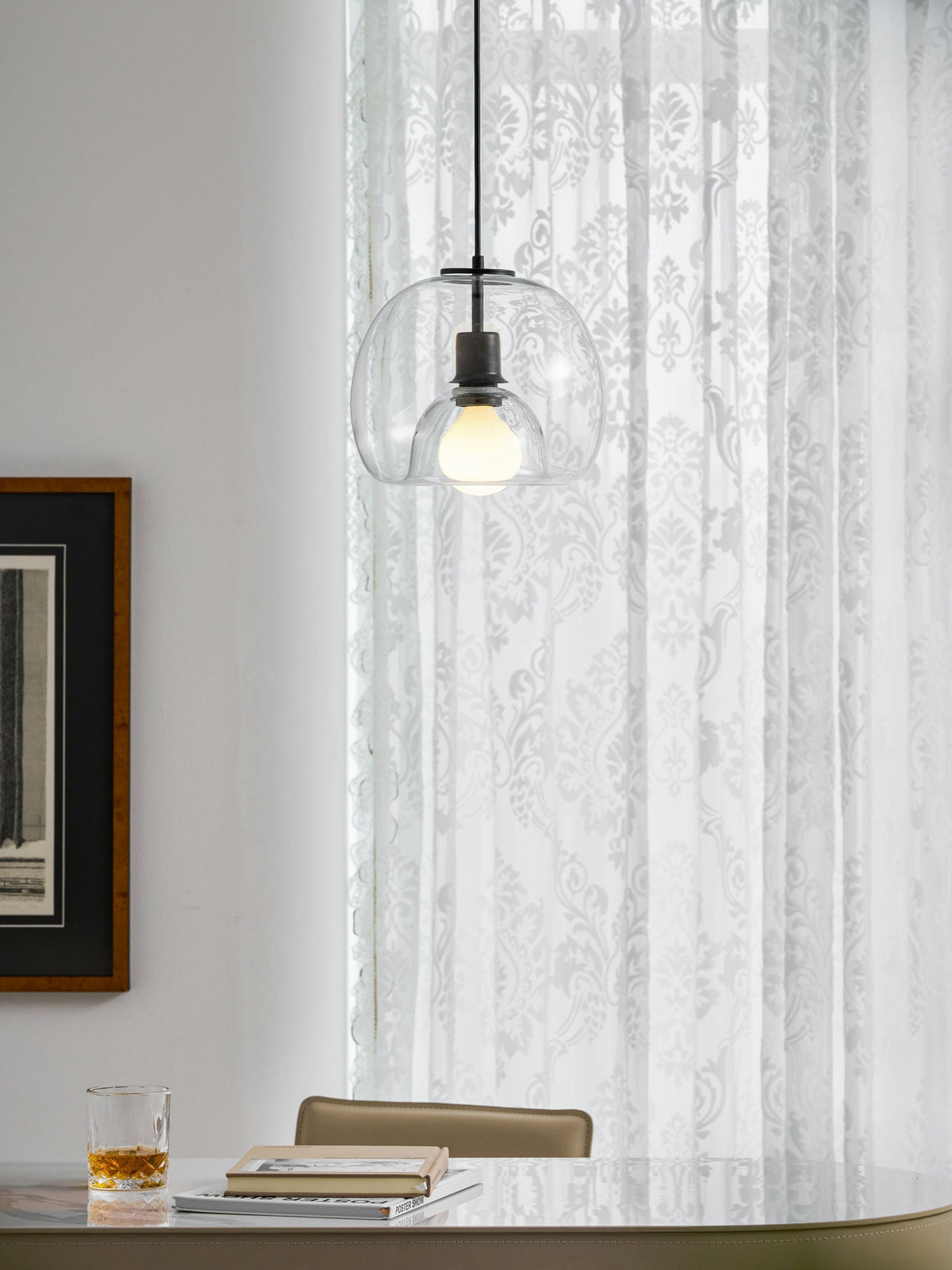 A clear glass Clérachic pendant hangs above a stone table with a glass of whisky and an open book, beside a dark frame.