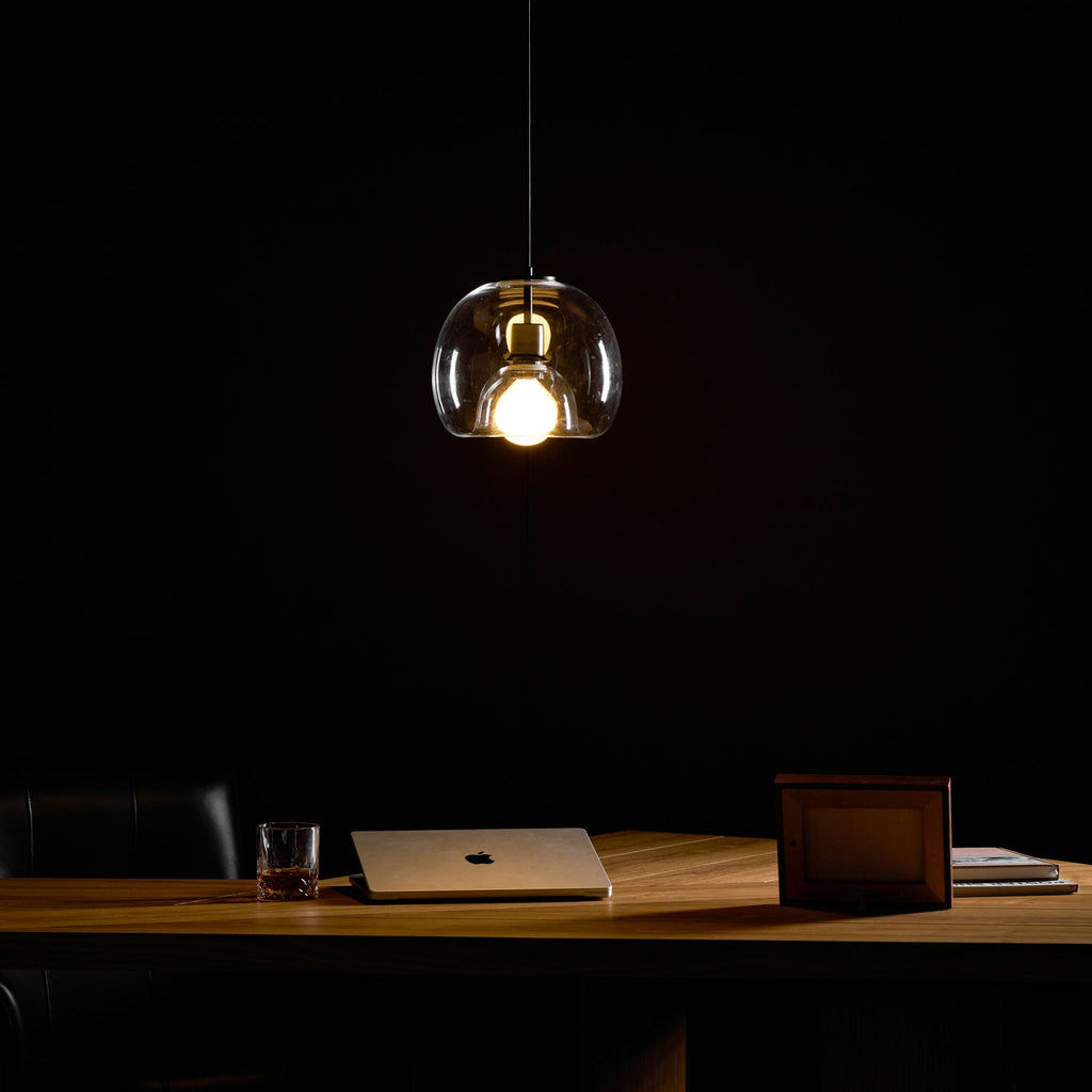A clear glass Clérachic pendant glows warm in a dark room, above a wood desk with a laptop and a framed photo.