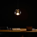 A clear glass Clérachic pendant glows warm in a dark room, above a wood desk with a laptop and a framed photo.