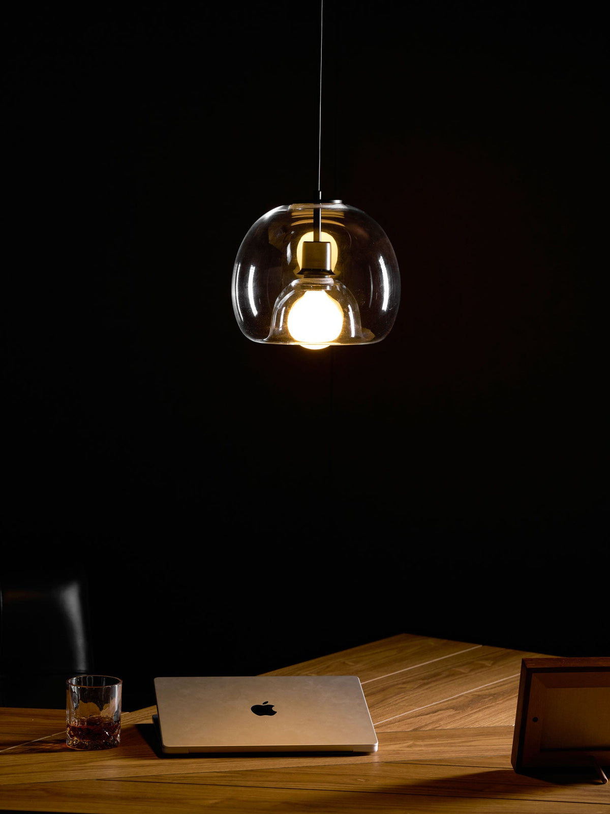 The same clear glass Clérachic pendant lit in a dark room, hung above a wood desk with a laptop and a whisky glass.