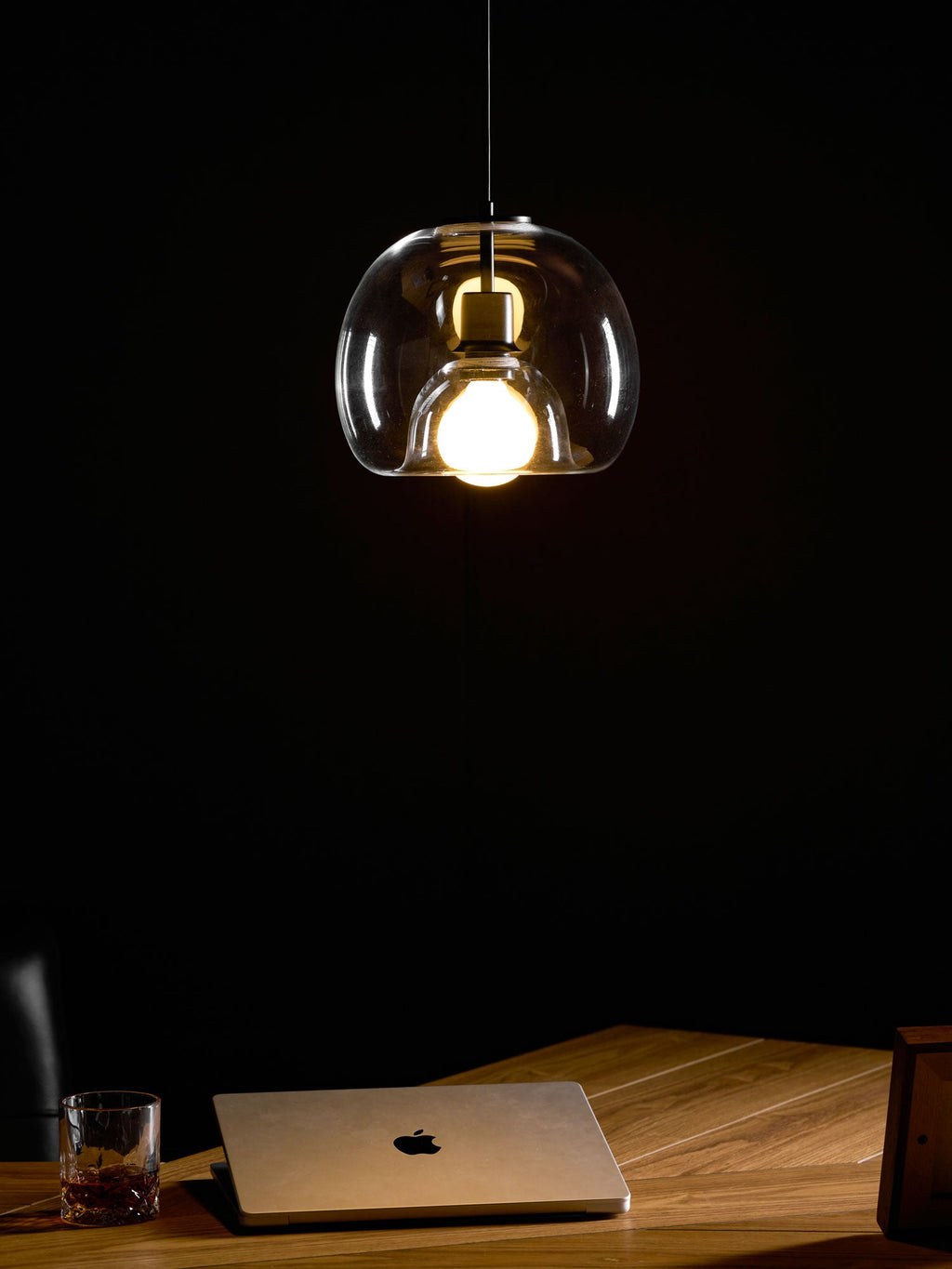 A lit clear glass Clérachic pendant seen in a dark room, with a laptop and glass on a wood desk below.