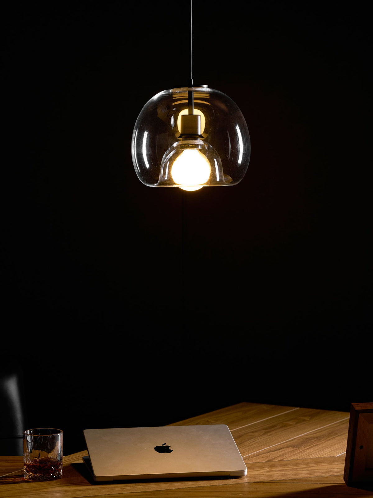 A lit clear glass Clérachic pendant seen in a dark room, with a laptop and glass on a wood desk below.