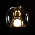 Close view of this Clérachic pendant lit in the dark, showing the clear outer shade, brass-toned socket and frosted bulb.