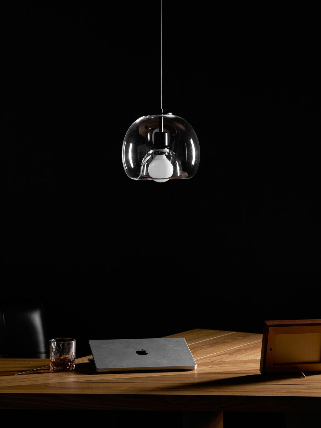 An unlit clear glass Clérachic pendant hangs in a dark room above a wood desk with a laptop and a whisky glass.