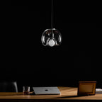 A Clérachic pendant in clear glass hangs unlit against a dark backdrop, with a framed photo and laptop on the desk.