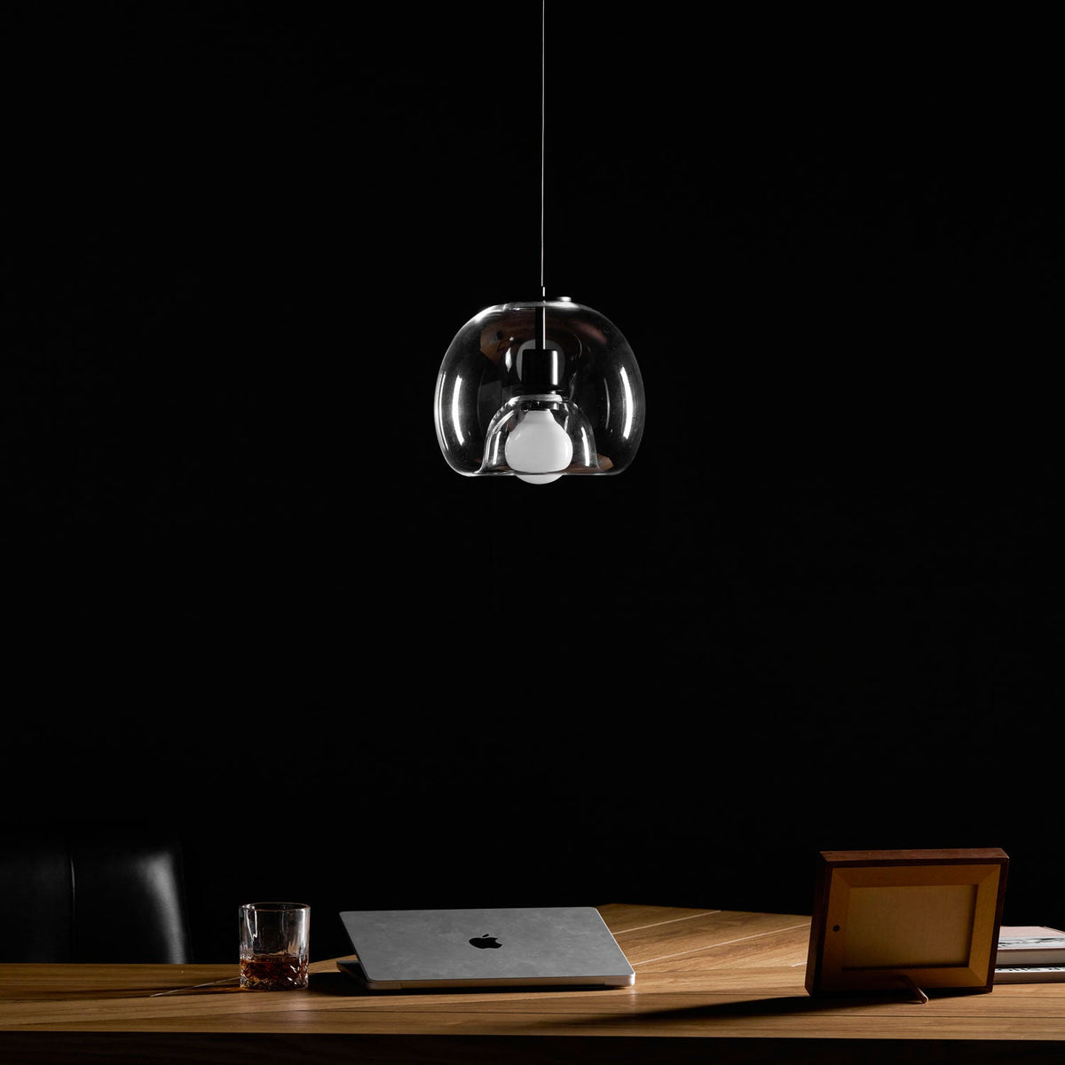 A Clérachic pendant in clear glass hangs unlit against a dark backdrop, with a framed photo and laptop on the desk.