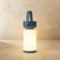 Clérachic cordless cylinder lamp glowing warm white, with a blue handle and base, on a limewashed plaster backdrop.