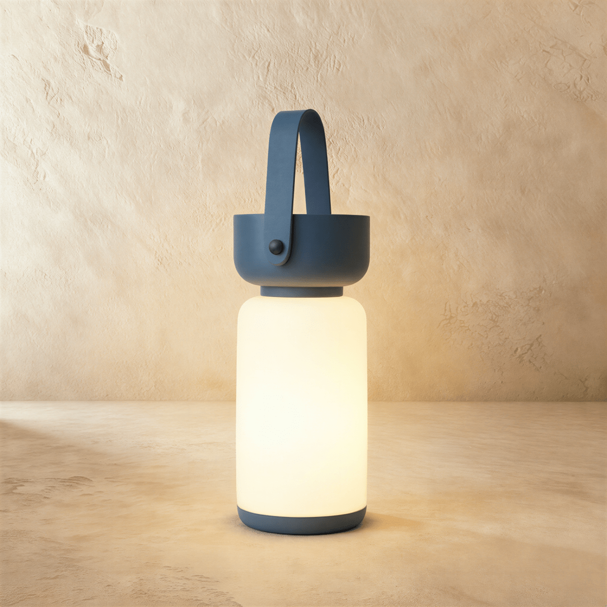 Clérachic cordless cylinder lamp glowing warm white, with a blue handle and base, on a limewashed plaster backdrop.