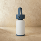Clérachic cordless cylinder lamp switched off, a white frosted body with a blue handle and base against plaster.