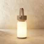 Clérachic cordless cylinder lamp lit warm white, with a taupe-brown handle and base on a limewashed plaster backdrop.