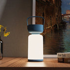 Clérachic cordless cylinder lamp with a blue handle glowing on a wooden shelf by yellow flowers and a canal painting.