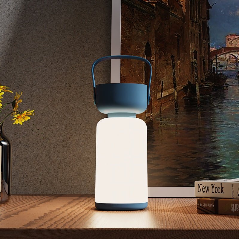 Clérachic cordless cylinder lamp with a blue handle glowing on a wooden shelf by yellow flowers and a canal painting.
