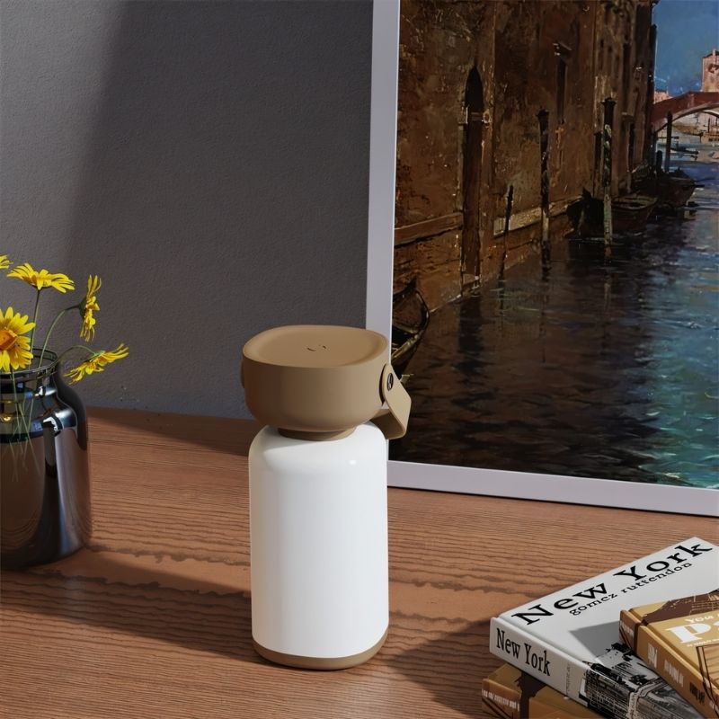 Clérachic cordless cylinder lamp, switched off with its brown handle tipped aside, on a wooden desk by a vase and books.